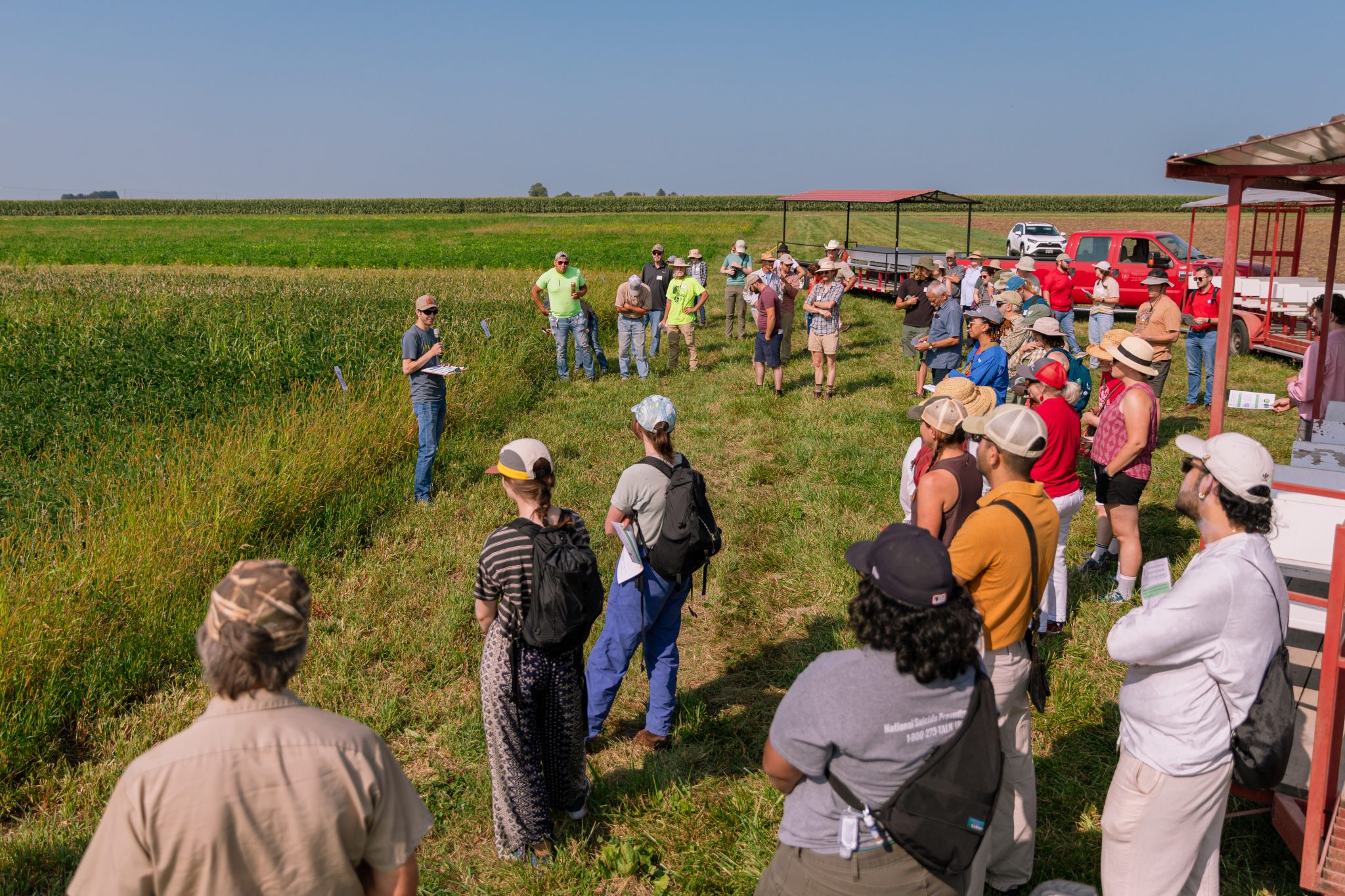 A researcher stands at the edge of a field speaking to a crowd of people.