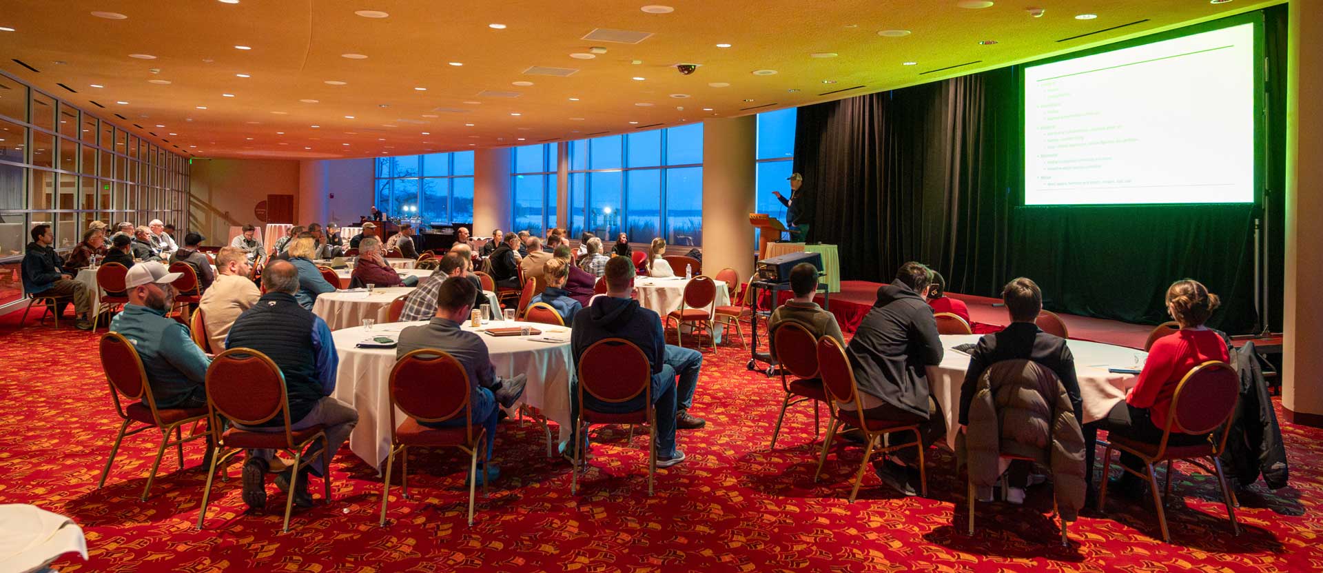 Attendees sit at round tables while listening to a speaker on stage.